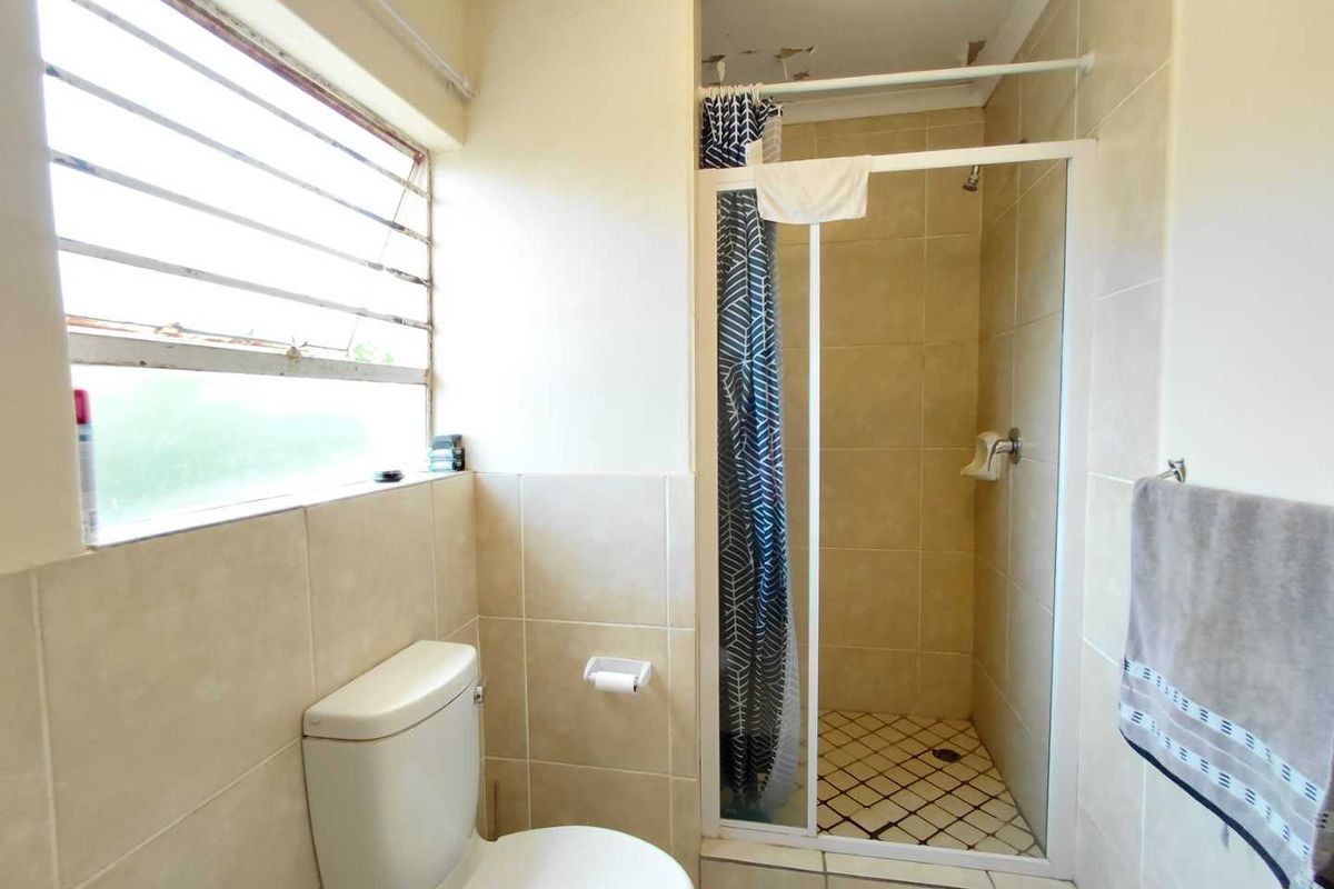 A functional bathroom featuring a tiled shower enclosure, standard toilet, and a large window with security bars.