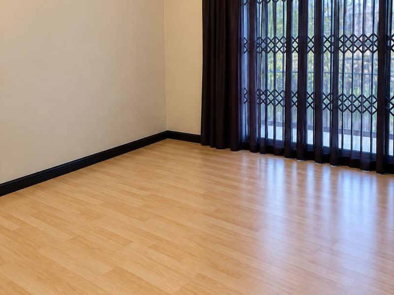 A bright, empty living room featuring light wood-look laminate flooring, neutral walls, and a sliding glass door with a security gate and dark curtains.
