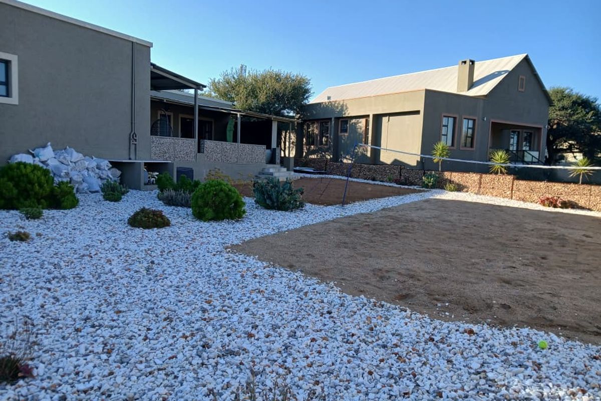 Spacious rear exterior view featuring a landscaped garden with gravel beds, a sand court, and a covered patio area attached to the main dwelling.