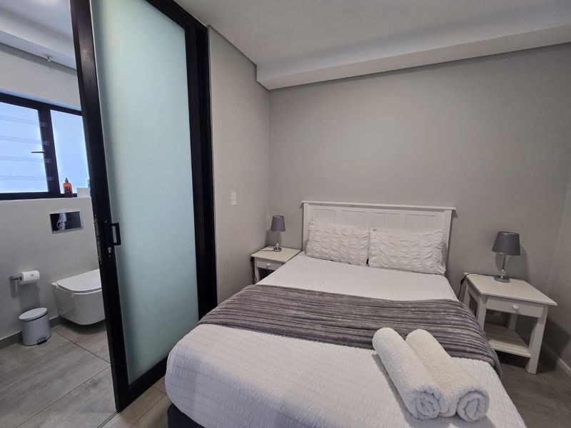 A modern bedroom featuring a sleek frosted glass sliding door leading to an en-suite bathroom with a wall-mounted toilet and tiled finishes.