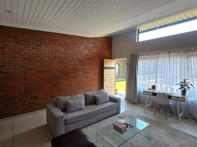 Spacious open-plan living area featuring a striking exposed brick wall, high sloped ceilings, and large windows that provide ample natural light.