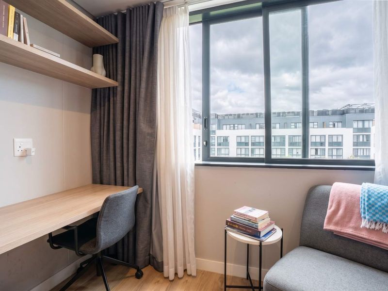 A modern, well-lit home office featuring a built-in desk, floating shelving, and large sliding windows with views of the surrounding apartment complex.