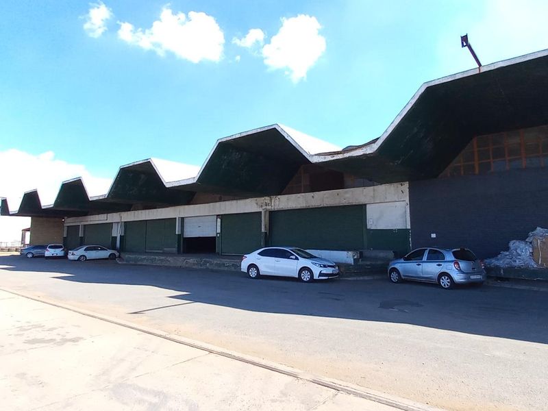 Industrial warehouse facility featuring a distinctive sawtooth roof design, multiple loading bays with roller shutter doors, and a large paved yard area.