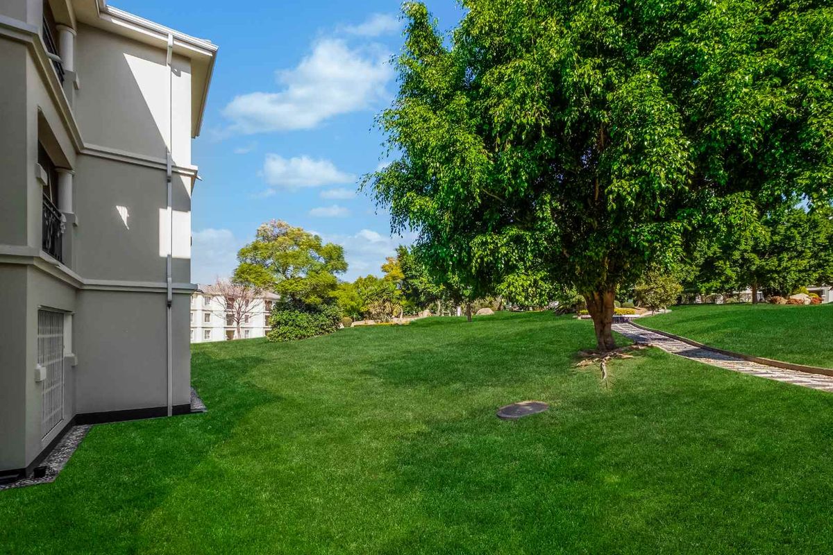 A well-maintained rear garden area featuring a lush green lawn, mature trees, and a paved pathway adjacent to the apartment building exterior.