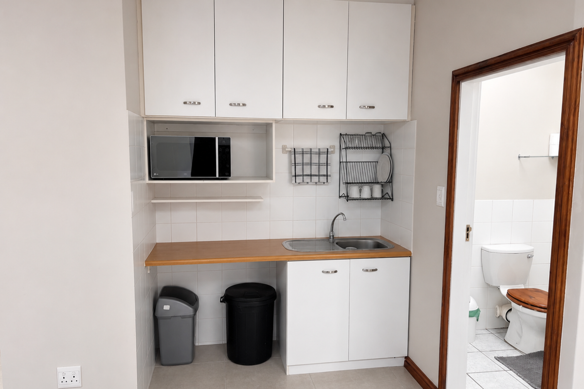 A compact office kitchenette featuring white cabinetry, a laminate countertop, stainless steel sink, and tiled backsplash, with an adjacent bathroom entrance.