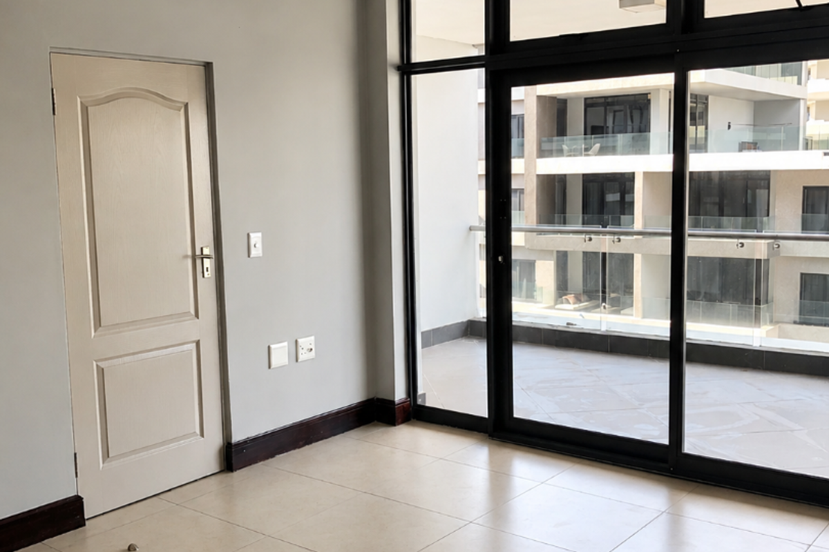 A bright, empty living area featuring neutral tiled flooring, modern grey walls, and large sliding glass doors leading to a private balcony.