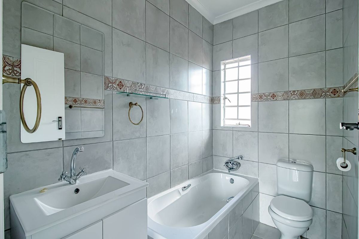 A clean, tiled bathroom featuring a built-in bathtub, a modern vanity with a basin, a toilet, and a window for natural light.