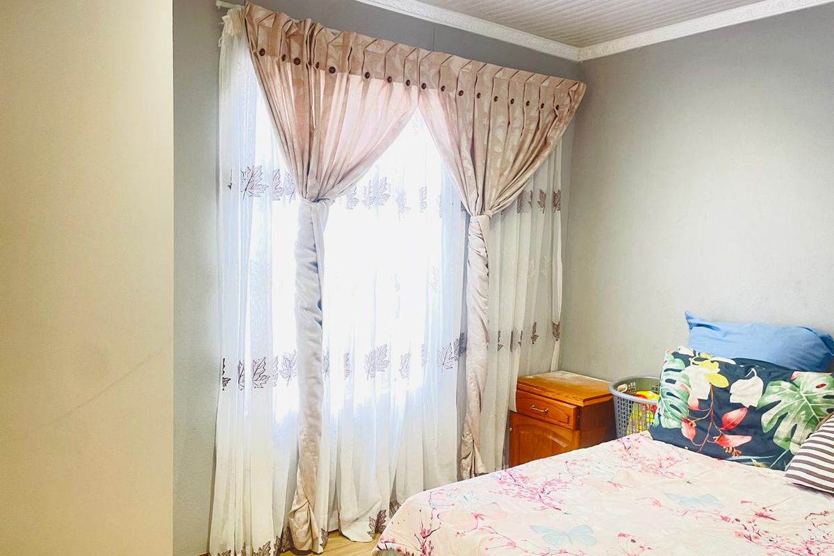A bedroom featuring light-toned laminate flooring, decorative ceiling cornicing, and a large window dressed with layered curtains.