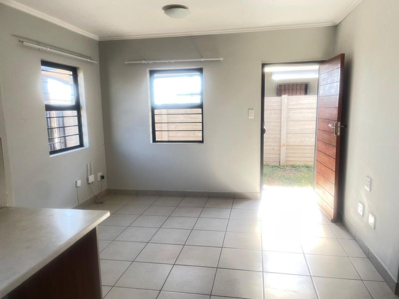 An empty open-plan living area featuring tiled flooring, security bars on windows, and direct access to the exterior through a timber door.