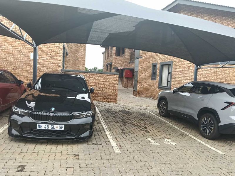 A paved outdoor parking area featuring a shade cloth carport structure set against a facebrick townhouse building.
