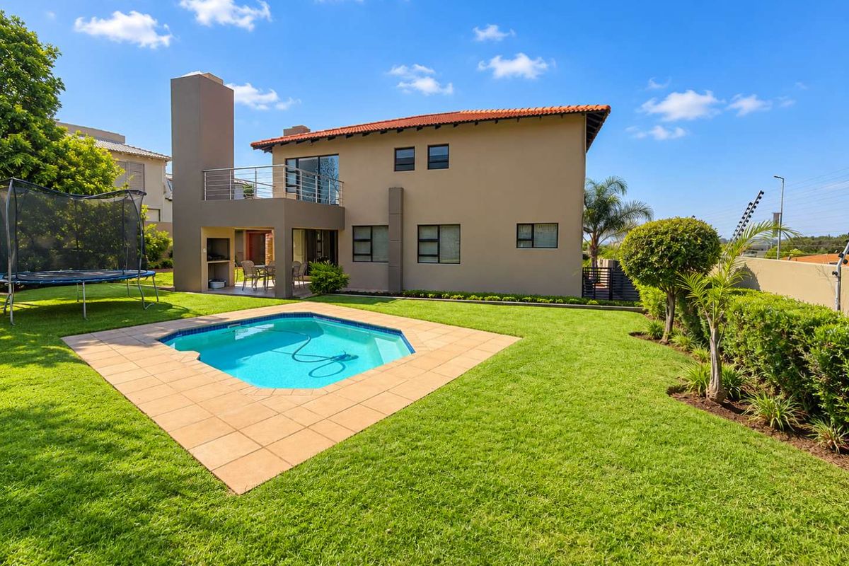 Rear view of the property featuring a well-maintained garden, swimming pool with tiled surround, covered patio with built-in braai, and an upper-level balcony.