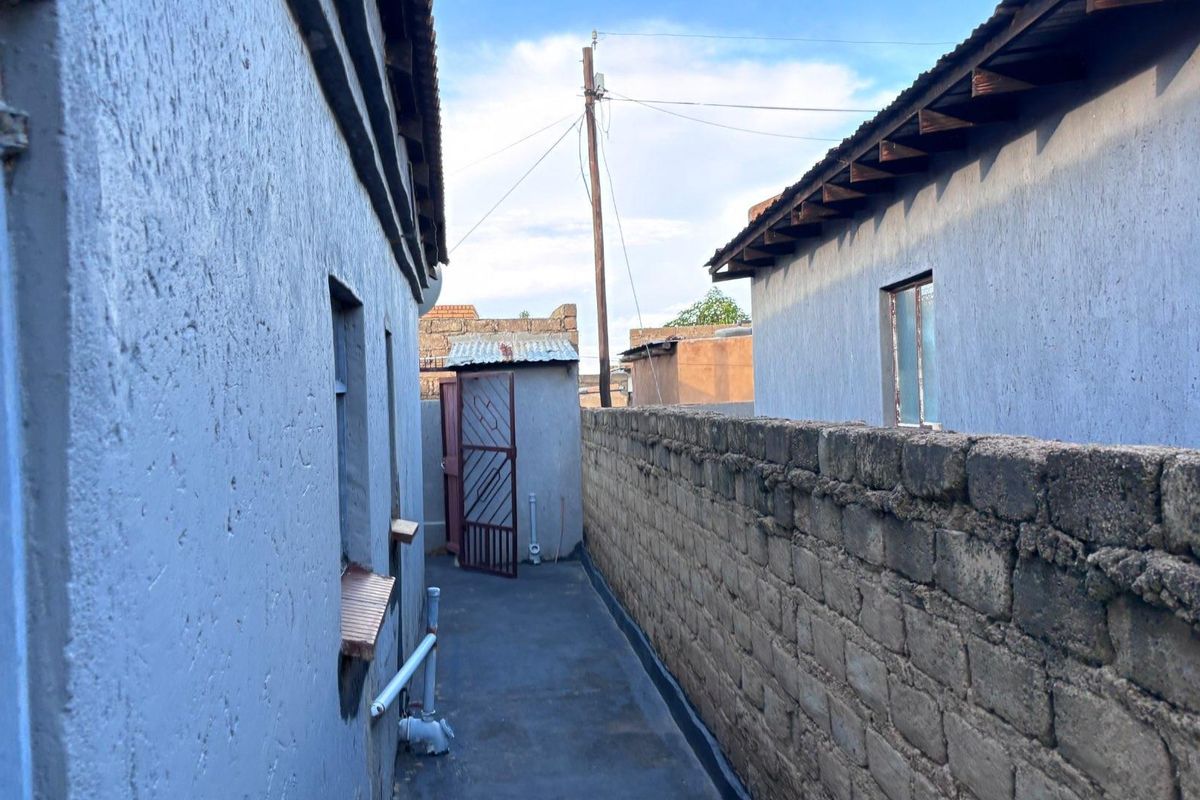 A narrow exterior side passage between two structures, featuring textured walls, a concrete walkway, and a metal gate leading to the rear area.