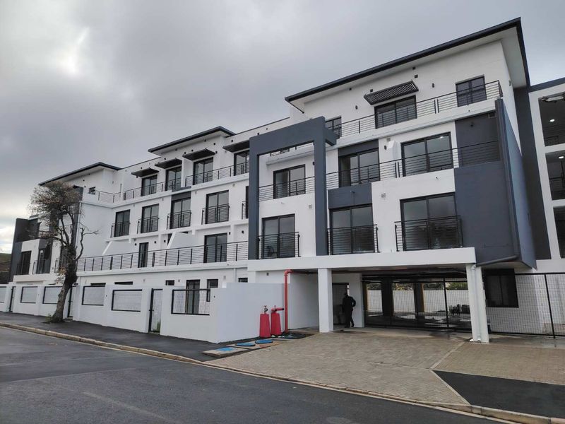 Modern multi-story apartment complex featuring a clean white and grey facade, private balconies with metal railings, and a paved ground-level entrance area.