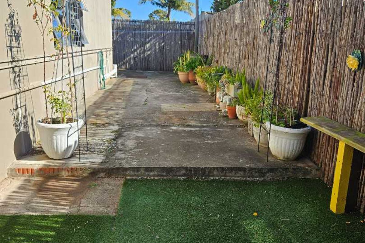 A rear outdoor area featuring a concrete pathway, artificial turf, and a metal garden arch, enclosed by reed fencing.