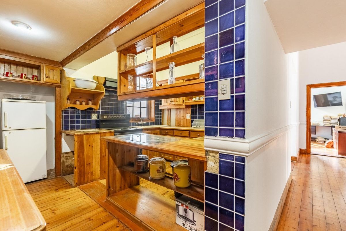 A rustic kitchen featuring wooden cabinetry, blue tiled backsplash, and wooden countertops, with an adjacent view into an office space.