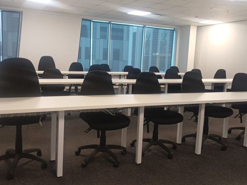 A functional office training room featuring large windows, suspended ceilings with recessed lighting, and carpeted flooring, set up with multiple desk stations.