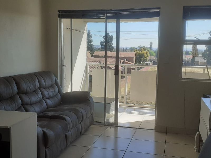 A living room featuring tiled flooring, large sliding glass doors leading to a balcony, and natural light through aluminium-framed windows.