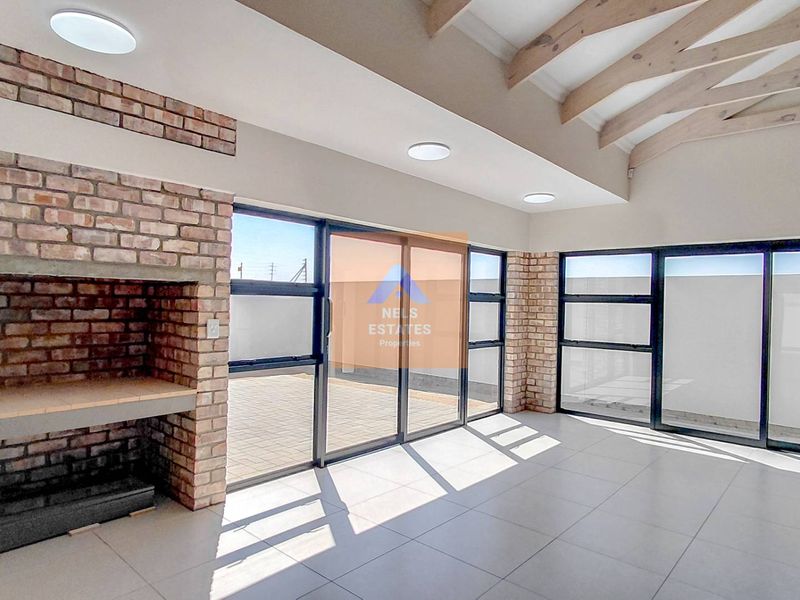 Spacious covered patio featuring a built-in brick fireplace, exposed timber ceiling beams, tiled flooring, and large aluminium sliding doors leading to the exterior.