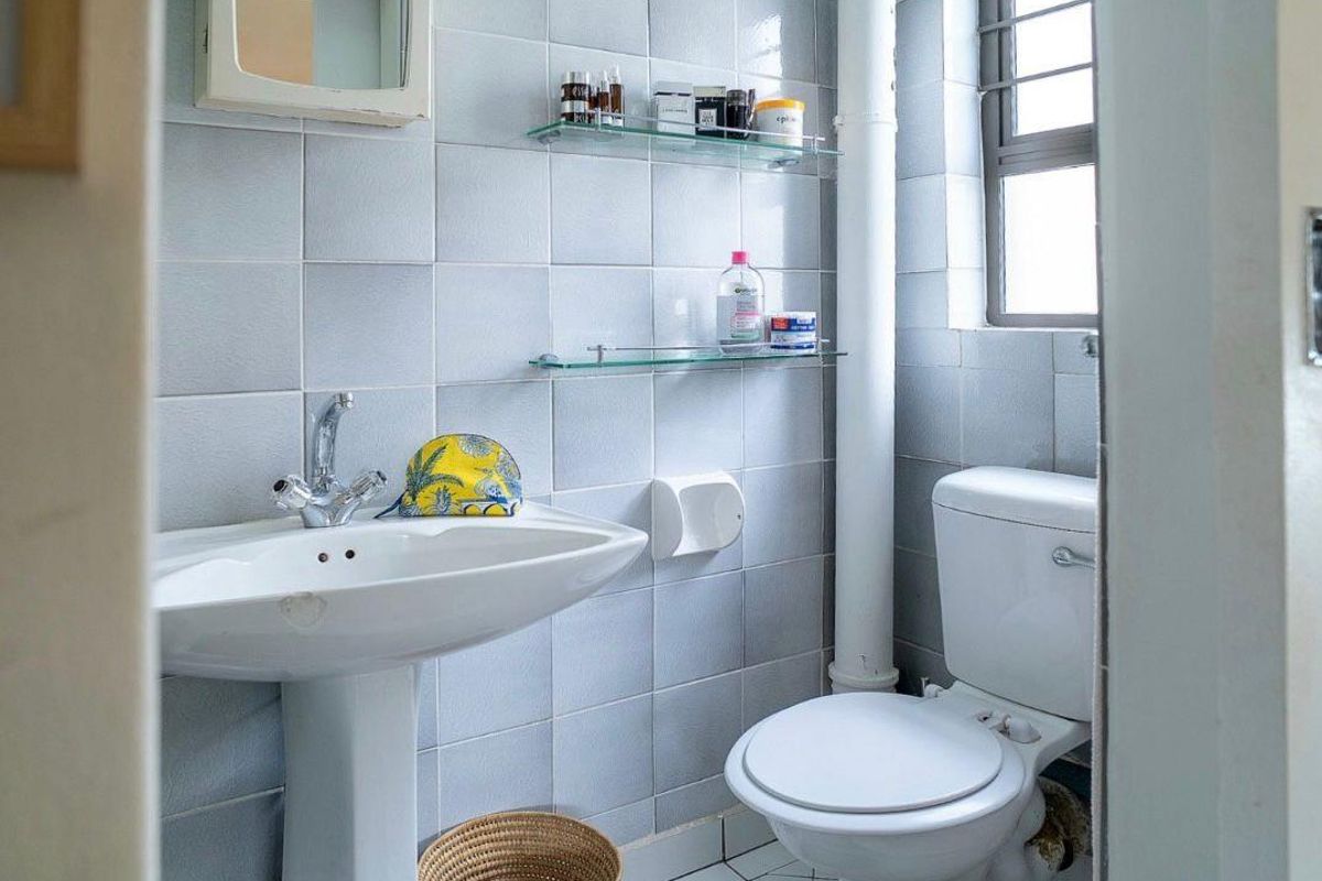 A compact bathroom featuring a pedestal basin, standard toilet, and light blue wall tiling with a window for natural ventilation.