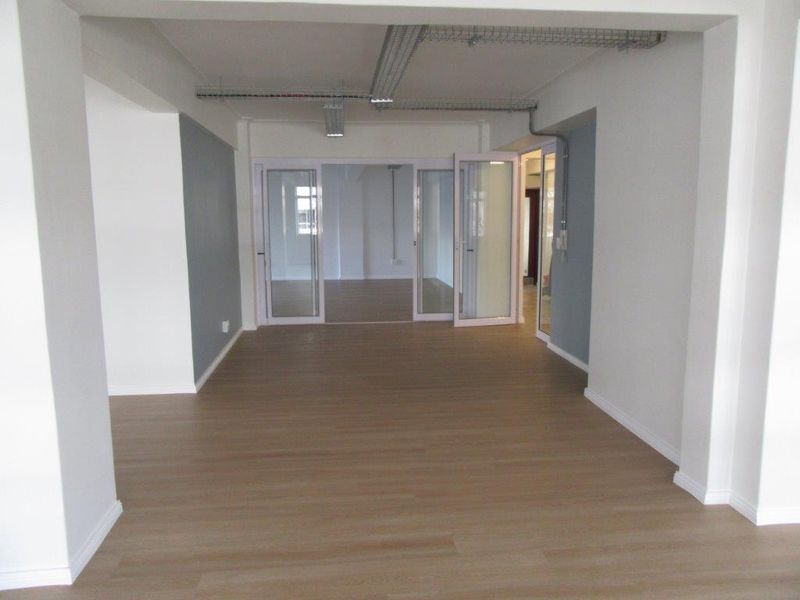 A bright, open-plan office or commercial space featuring modern laminate flooring, exposed industrial-style cable management, and glass-paneled interior doors.