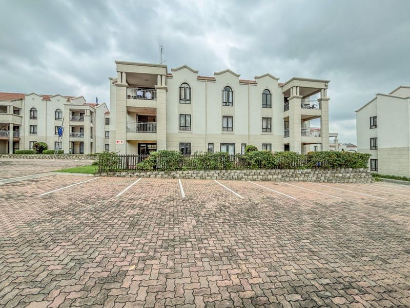 Exterior view of a multi-story apartment complex featuring paved parking area, balconies, and landscaped garden borders.