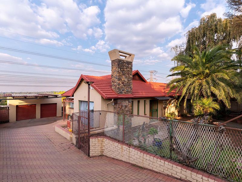 Front view of a residential property featuring a stone chimney, tiled roof, paved driveway, and a double garage with a brick and metal boundary wall.
