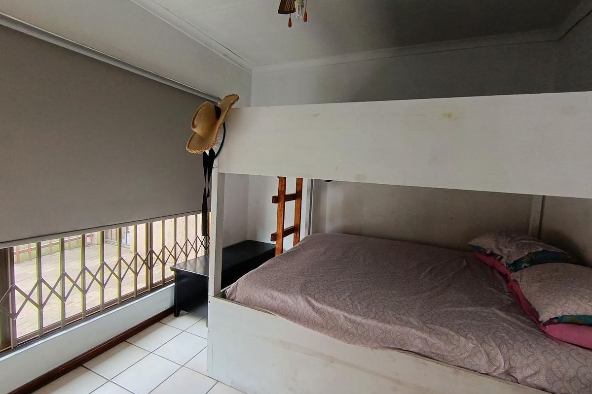 A bedroom featuring tiled flooring, a ceiling fan, and a window fitted with a roller blind and security gate.