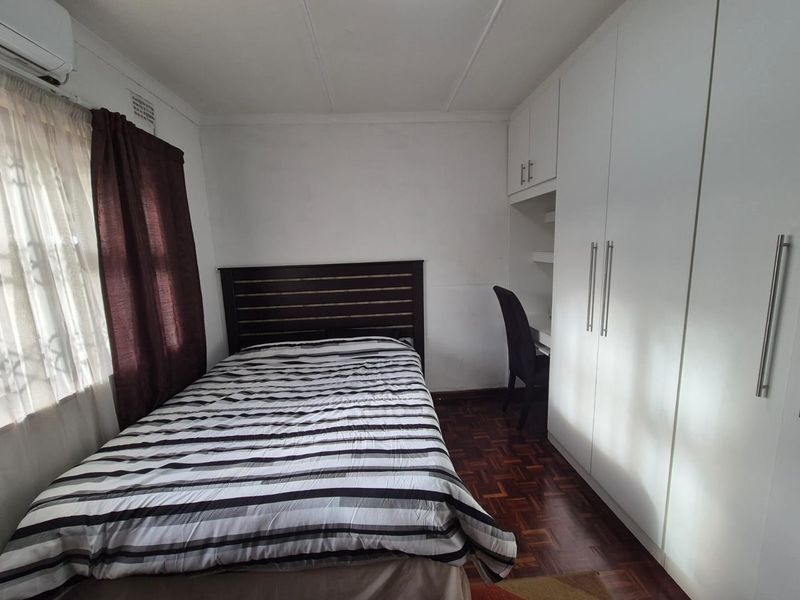 A functional bedroom featuring parquet flooring, built-in white cabinetry with an integrated desk space, and a wall-mounted air conditioning unit.