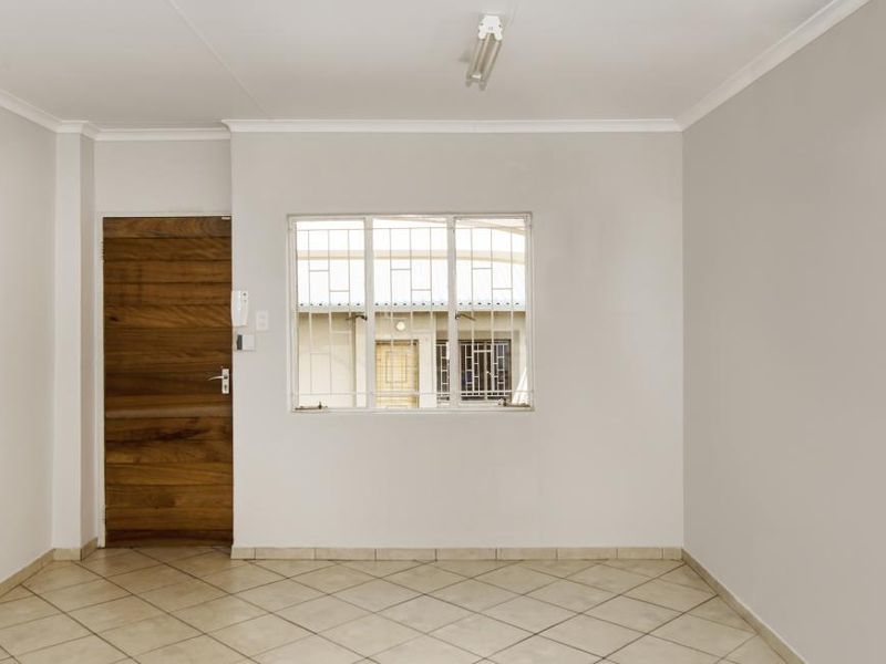 An empty, neutrally painted living area featuring tiled flooring, a wooden door, and a window fitted with security bars.