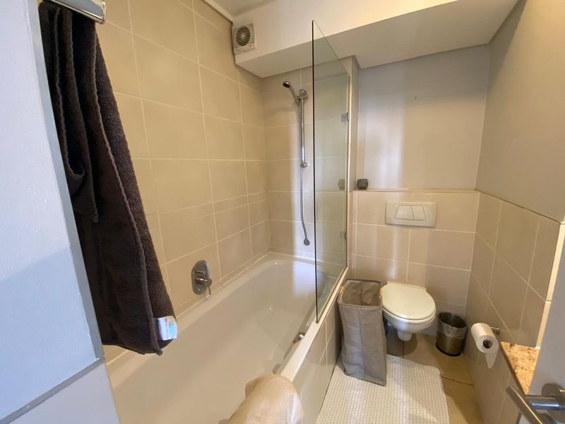 A functional bathroom featuring a bathtub with a glass shower screen, wall-hung toilet with concealed cistern, and neutral wall tiling.
