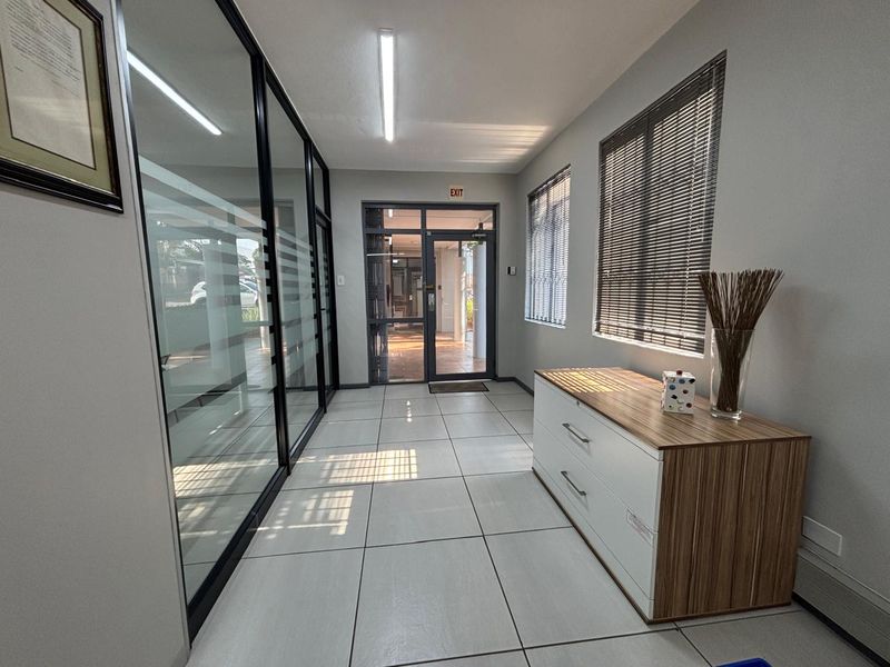 A bright office hallway featuring tiled flooring, glass partition walls, and an aluminium-framed glass entrance door with natural light.