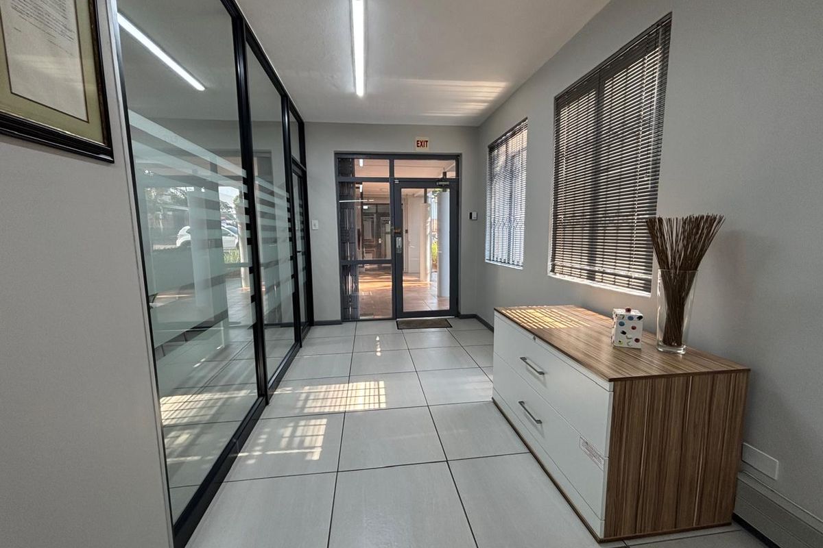 A bright office hallway featuring tiled flooring, glass partition walls, and an aluminium-framed glass entrance door with natural light.