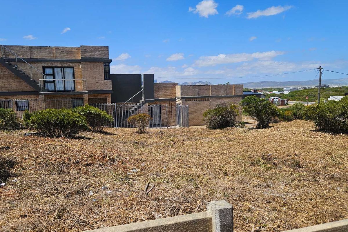 A view of a vacant residential plot with established shrubs, adjacent to a multi-level brick residential building with external staircases.