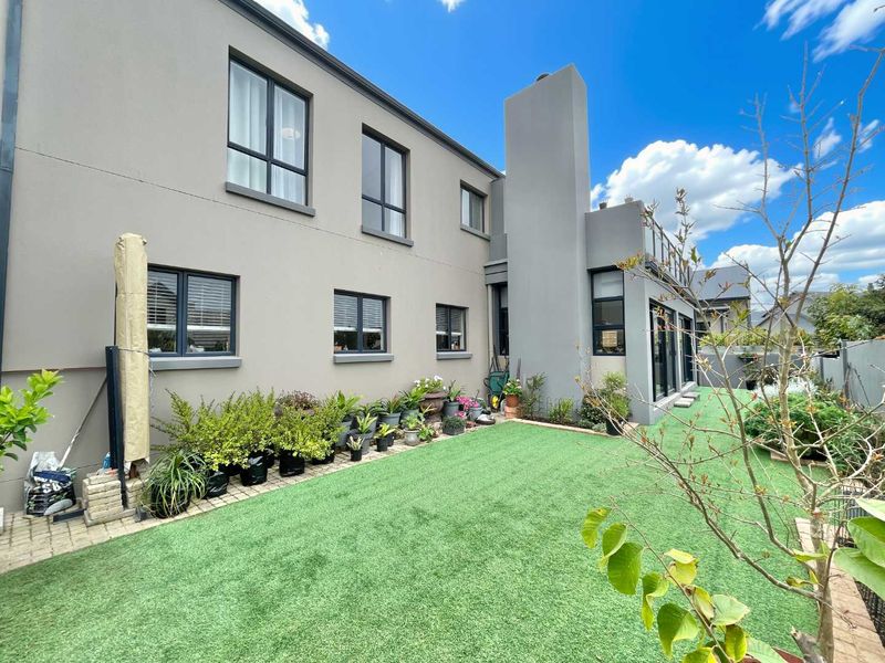 Rear view of a modern two-story house featuring a grey facade, artificial lawn, paved garden path, and upper-level balcony.