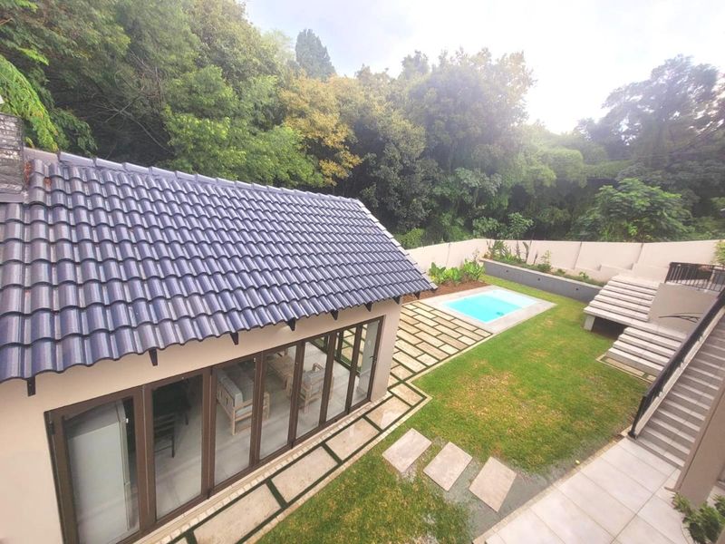 An elevated view of a rear garden featuring a swimming pool, paved pathways, and a modern structure with large sliding glass doors.