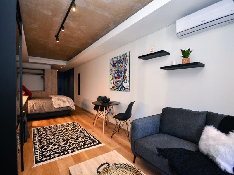 Modern open-plan studio apartment featuring exposed concrete ceilings, timber flooring, track lighting, and a wall-mounted split-system air conditioner.