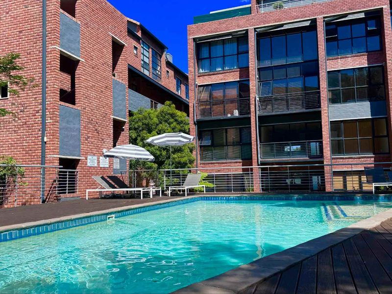 A well-maintained communal swimming pool area featuring timber decking, stainless steel railings, and a modern red brick apartment building facade.