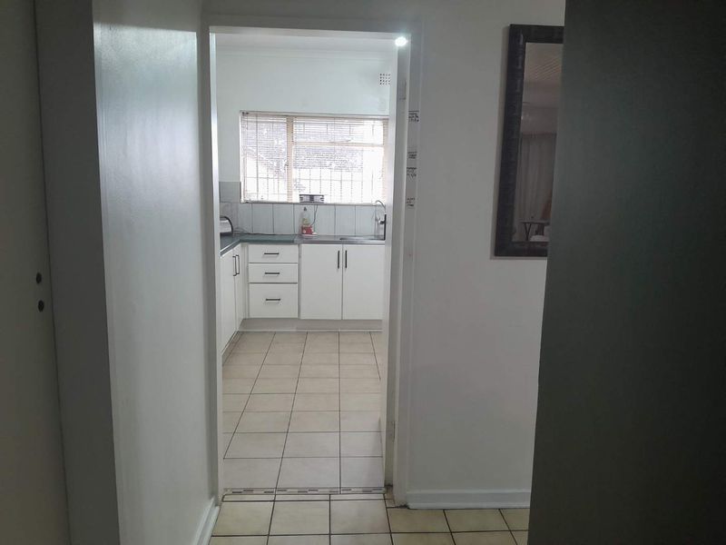 A functional kitchen featuring white cabinetry, tiled flooring, and a window with security bars, viewed from an adjacent hallway.