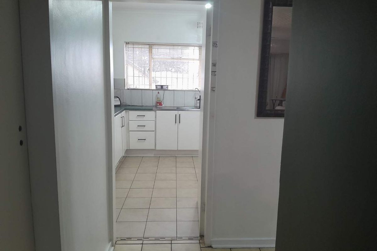 A functional kitchen featuring white cabinetry, tiled flooring, and a window with security bars, viewed from an adjacent hallway.