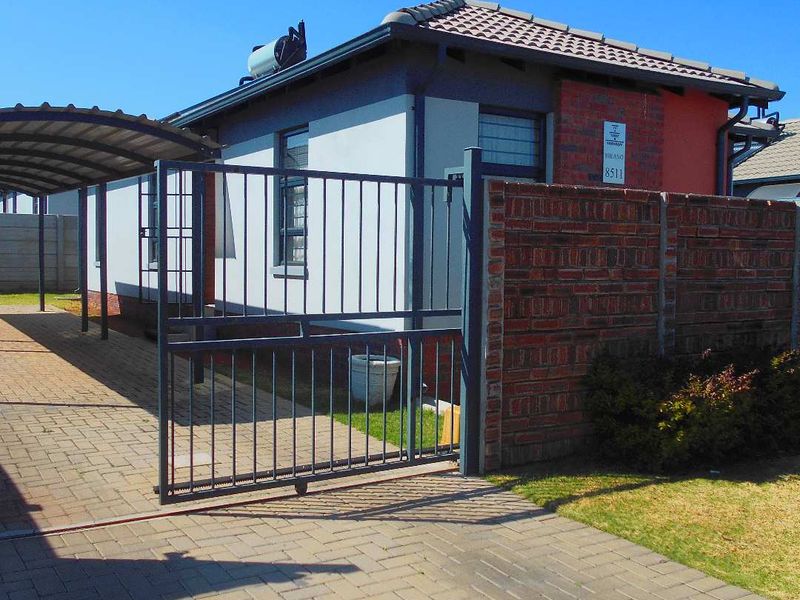 Front exterior view of a residential property featuring a paved driveway, carport, metal sliding gate, and brick boundary wall.