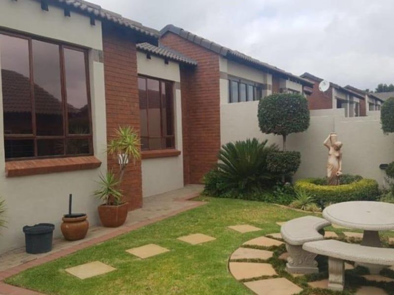 A well-maintained rear garden area featuring a brick facade, paved pathways, and established landscaping with a concrete seating area.