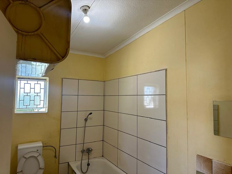 A dated bathroom featuring a bathtub with a handheld shower head, tiled splashback, and security window grille, requiring cosmetic updates and maintenance.