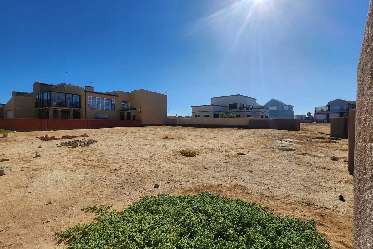 A vacant residential plot of land featuring sandy terrain, surrounded by established residential properties and boundary walls.