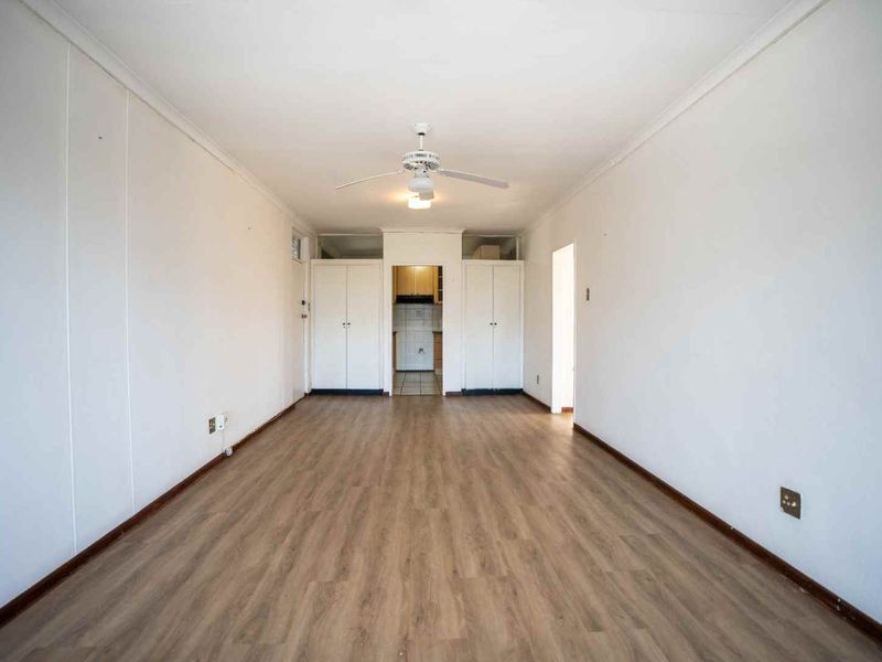 A spacious, empty living area featuring light wood-look laminate flooring, a ceiling fan, and built-in storage cupboards, with a view through to the kitchen.