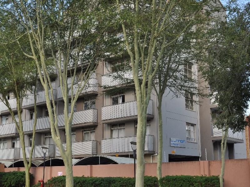 Exterior view of a multi-story apartment building featuring private balconies, security fencing, and a perimeter wall, surrounded by mature trees.