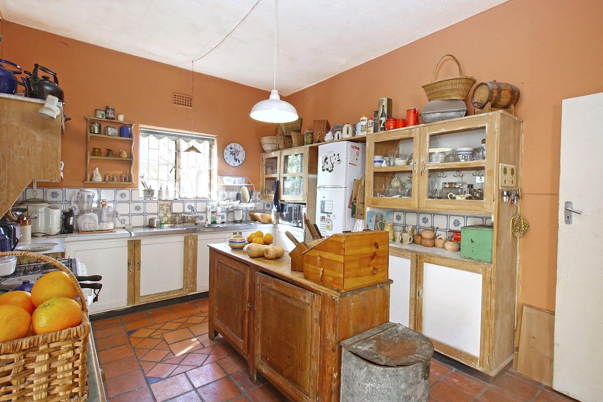 A rustic kitchen featuring terracotta floor tiles, built-in wooden cabinetry, a central island, and a tiled backsplash with a window overlooking the exterior.