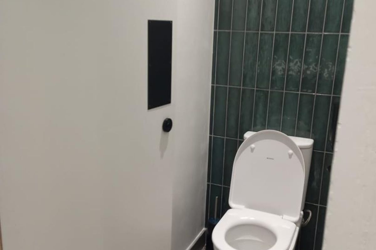 A clean, modern industrial-style restroom featuring a white toilet set against a dark green vertical tiled feature wall and grey floor tiles.