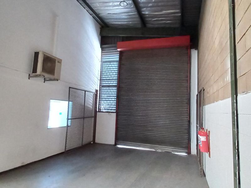 Industrial warehouse space featuring a high-clearance roller shutter door, exposed ceiling structure, concrete flooring, and wall-mounted air conditioning unit.