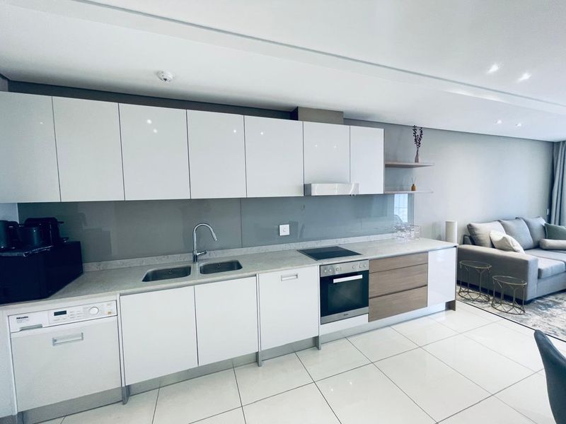 Modern open-plan kitchen featuring sleek white cabinetry, stone countertops, integrated appliances, and a glass splashback, seamlessly flowing into the living area.