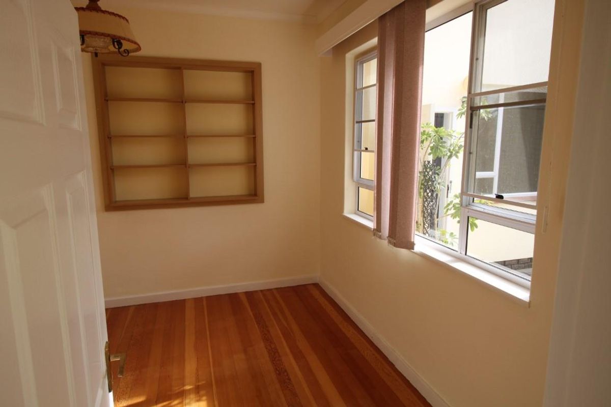 A bright, empty bedroom featuring polished timber flooring, built-in wall shelving, and large windows overlooking an exterior courtyard area.